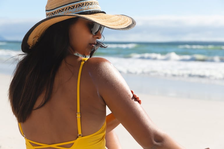 mujer con sombrero en la playa aplicando protector solar en la piel de su brazo